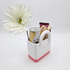 以下が含まれることがあります： 「Mami-Bag」の文字が入った白とピンクのトートバッグには、MALの飲み物の缶、スナックのパック、ストローが入っています。大きな白い花がトートバッグの横に置かれています。