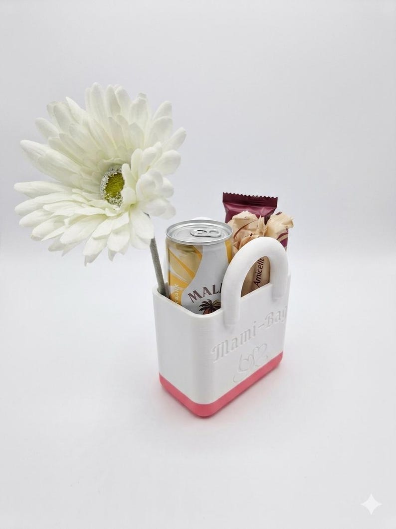 以下が含まれることがあります： 「Mami-Bag」の文字が入った白とピンクのトートバッグには、MALの飲み物の缶、スナックのパック、ストローが入っています。大きな白い花がトートバッグの横に置かれています。