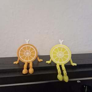May include: Two whimsical fruit slice figurines, one orange and one lemon, each with arms, legs, and a small white stem. The orange slice is orange with white segments, and the lemon slice is yellow with white segments.