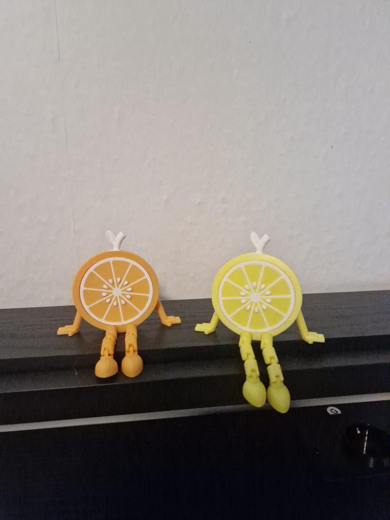 May include: Two whimsical fruit slice figurines, one orange and one lemon, each with arms, legs, and a small white stem. The orange slice is orange with white segments, and the lemon slice is yellow with white segments.