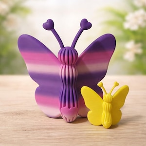 May include: Two butterfly-shaped ornaments. The larger one has purple and pink striped wings and a detailed body. The smaller one is yellow. Both have heart-shaped antennae. They are on a light wood surface.