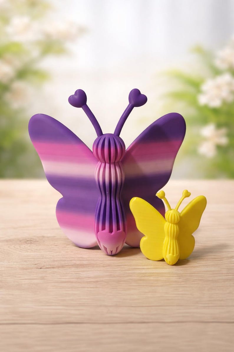 May include: Two butterfly-shaped ornaments. The larger one has purple and pink striped wings and a detailed body. The smaller one is yellow. Both have heart-shaped antennae. They are on a light wood surface.