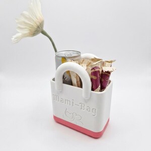 以下が含まれることがあります： ピンクのベースとハンドルが付いた白いトートバッグ。缶、スナック、白い花が入っています。バッグには「Mami-Bag」の文字とハートのデザインがあります。花は白い花びらと緑色の茎をしています。