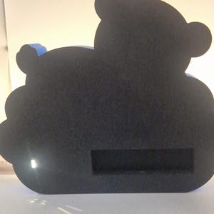 Puede incluir: Un objeto decorativo negro con forma de oso con una ranura rectangular. El oso tiene dos orejas peque&ntilde;as y un acento azul en la espalda. El art&iacute;culo est&aacute; hecho de un material texturizado.