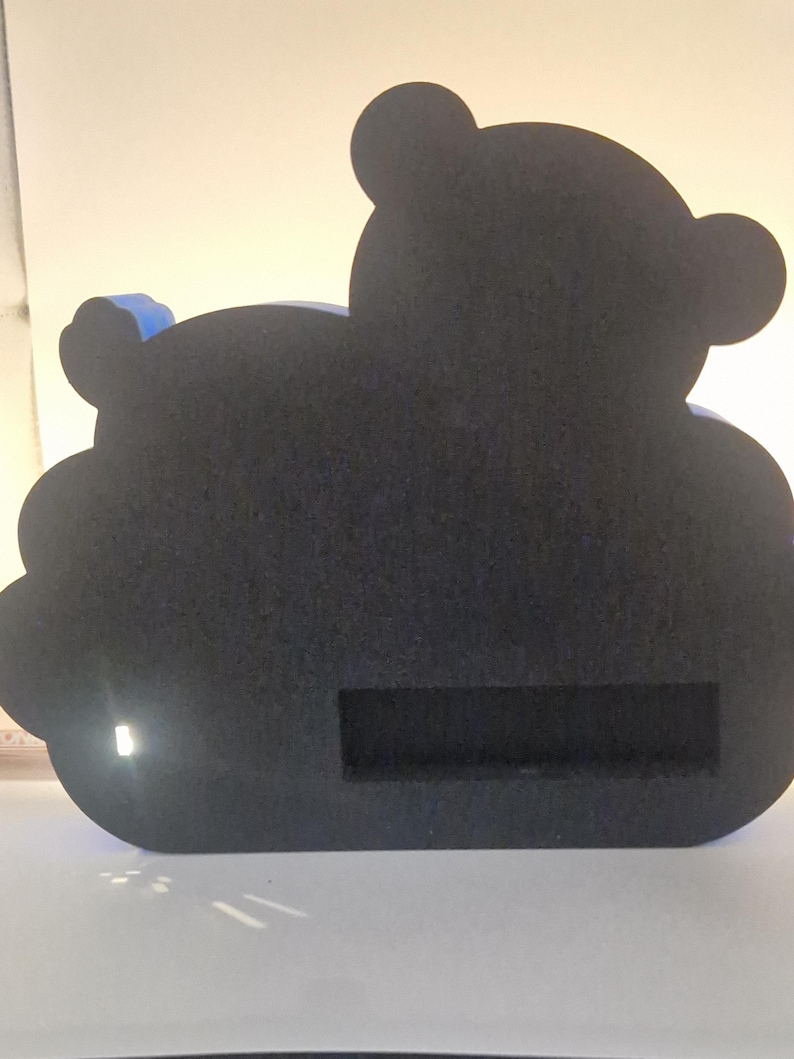 Puede incluir: Un objeto decorativo negro con forma de oso con una ranura rectangular. El oso tiene dos orejas peque&ntilde;as y un acento azul en la espalda. El art&iacute;culo est&aacute; hecho de un material texturizado.