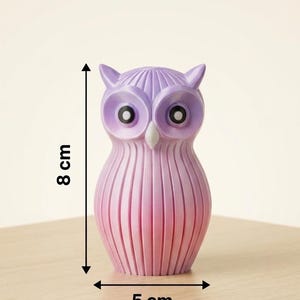 Puede incluir: Una figura decorativa de b&uacute;ho, de aproximadamente 5 cm de ancho y 8 cm de alto, en tonos rosas y morados. El b&uacute;ho tiene ojos grandes y un cuerpo texturizado y acanalado. Se exhibe sobre una superficie de madera.