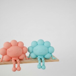 K&ouml;nnte beinhalten: Zwei skurrile, bl&uuml;tenf&ouml;rmige Figuren in Koralle und Hellblau, jede mit einem l&auml;chelnden Gesicht und h&auml;ngenden Beinen, sitzen auf einem kleinen Holzregal. Die Figuren haben ein verspieltes Design, perfekt f&uuml;r ein Kinderzimmer oder als dekorativer Akzent.