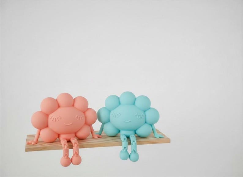 K&ouml;nnte beinhalten: Zwei skurrile, bl&uuml;tenf&ouml;rmige Figuren in Koralle und Hellblau, jede mit einem l&auml;chelnden Gesicht und h&auml;ngenden Beinen, sitzen auf einem kleinen Holzregal. Die Figuren haben ein verspieltes Design, perfekt f&uuml;r ein Kinderzimmer oder als dekorativer Akzent.