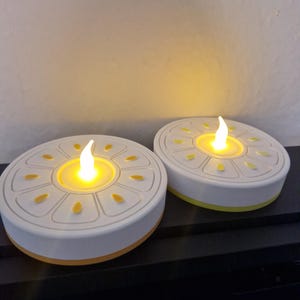 May include: Two battery-operated LED candles with a white base and yellow accents. The candles have a round shape with a decorative design on top and a flickering flame effect. The candles are designed to look like real candles.