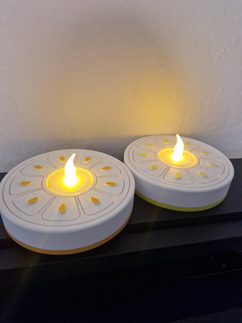 May include: Two battery-operated LED candles with a white base and yellow accents. The candles have a round shape with a decorative design on top and a flickering flame effect. The candles are designed to look like real candles.