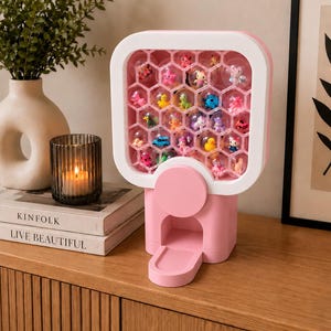 Könnte beinhalten: Ein rosa-weißer Verkaufsautomat aus Kunststoff mit einer wabenförmigen Gitterstruktur. Jede sechseckige Zelle enthält eine kleine, durchsichtige Kunststoffkugel mit einer bunten Miniaturfigur darin. Das Gerät hat ein rundes rosa Einstellrad.