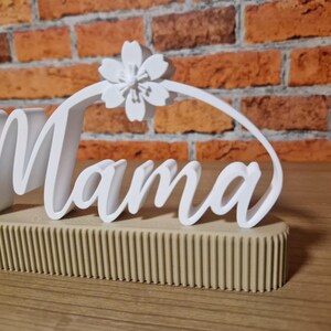 May include: A white 'Mama' sign with a floral detail, set on a beige, ribbed base. The sign is displayed against a brick wall background. The word 'Mama' is written in a cursive font.