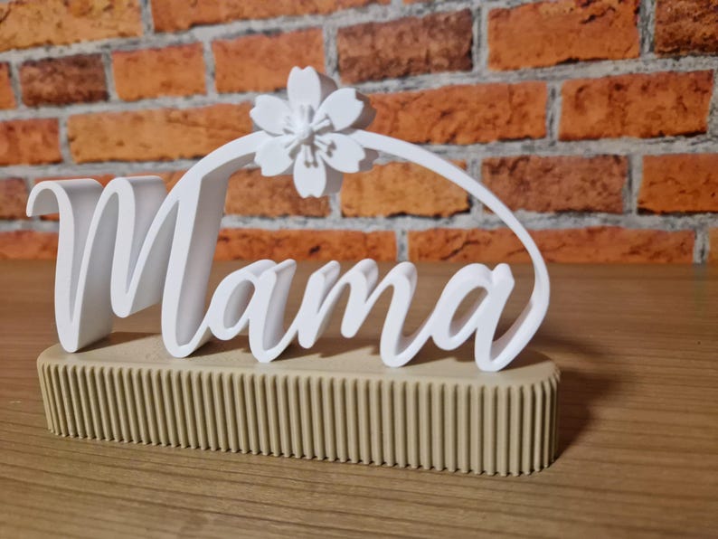 May include: A white 'Mama' sign with a floral detail, set on a beige, ribbed base. The sign is displayed against a brick wall background. The word 'Mama' is written in a cursive font.