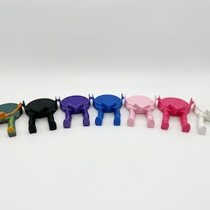 May include: A selection of colourful phone stands in a range of shades: green, black, purple, blue, pink, red, and white. Each stand features a circular top and two leg-like supports, creating a unique, artistic design.