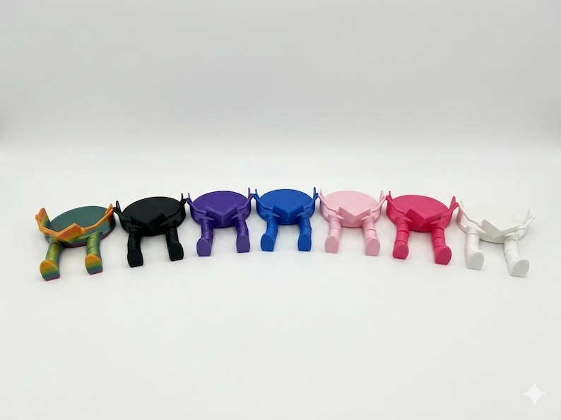 May include: A selection of colourful phone stands in a range of shades: green, black, purple, blue, pink, red, and white. Each stand features a circular top and two leg-like supports, creating a unique, artistic design.