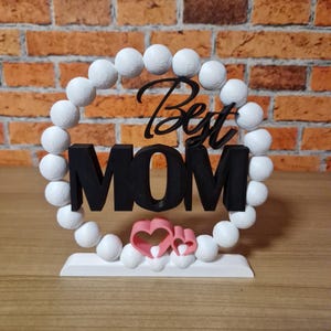 以下が含まれることがあります： 白いビーズで囲まれた黒い文字「Best MOM」と2つのピンクのハート。白いベースの上にデザインが配置されています。背景はレンガの壁です。