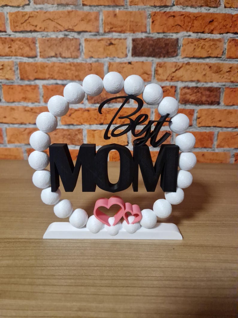 以下が含まれることがあります： 白いビーズで囲まれた黒い文字「Best MOM」と2つのピンクのハート。白いベースの上にデザインが配置されています。背景はレンガの壁です。
