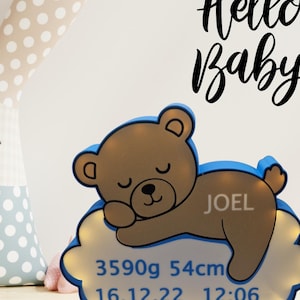 Puede incluir: Una luz de noche con forma de oso de peluche marr&oacute;n con borde azul, sobre una base en forma de nube. La base muestra el nombre "JOEL", peso 3590g, longitud 54cm y fecha de nacimiento 16.12.22 con hora 12:06. Las palabras "Hello Baby" est&aacute;n escritas arriba.