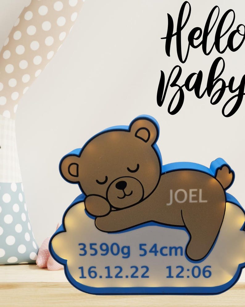 Puede incluir: Una luz de noche con forma de oso de peluche marr&oacute;n con borde azul, sobre una base en forma de nube. La base muestra el nombre "JOEL", peso 3590g, longitud 54cm y fecha de nacimiento 16.12.22 con hora 12:06. Las palabras "Hello Baby" est&aacute;n escritas arriba.