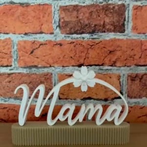 May include: A white 'Mama' sign in cursive script, featuring a floral embellishment, placed on a beige, textured base. The background is a brick wall.