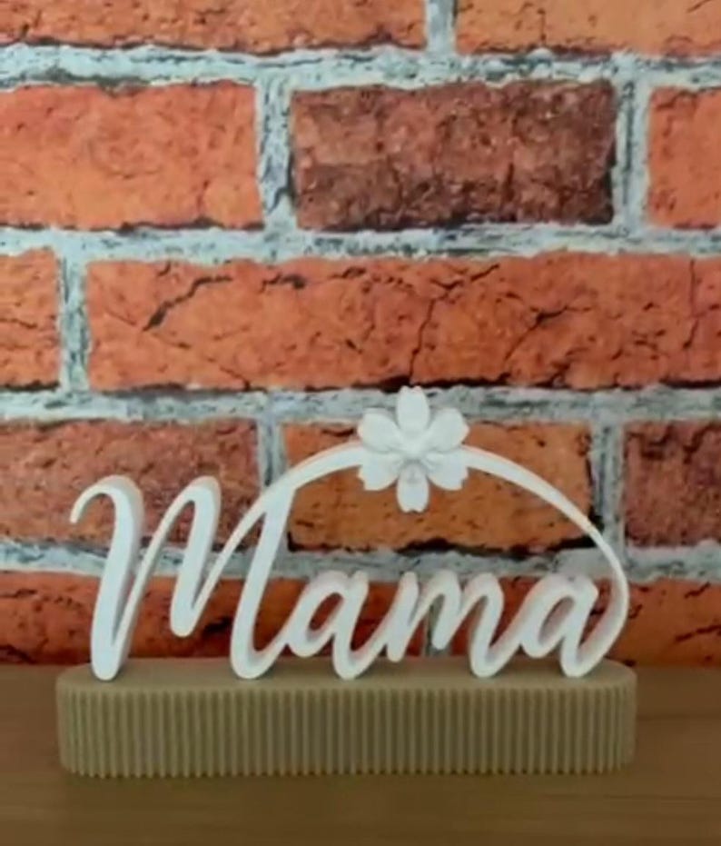 May include: A white 'Mama' sign in cursive script, featuring a floral embellishment, placed on a beige, textured base. The background is a brick wall.