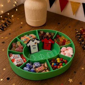 Könnte beinhalten: Ein grünes, ovales Snack-Tablett mit verschiedenen Fächern und einer weißen Fußballfeld-Grafik in der Mitte. Zwei kleine trikotförmige Marker liegen auf dem Tablett, ein weißer mit der Aufschrift JAMAL 10 und ein roter mit DAVID 8, gefüllt mit verschiedenen Süßigkeiten.