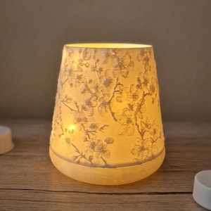 May include: A lit, cream-coloured candle holder with a floral pattern. The cylindrical holder has a flared top and a rounded base. The floral design is embossed on the surface, and the light from the candle inside illuminates the design. The candle holder sits on a wooden surface.