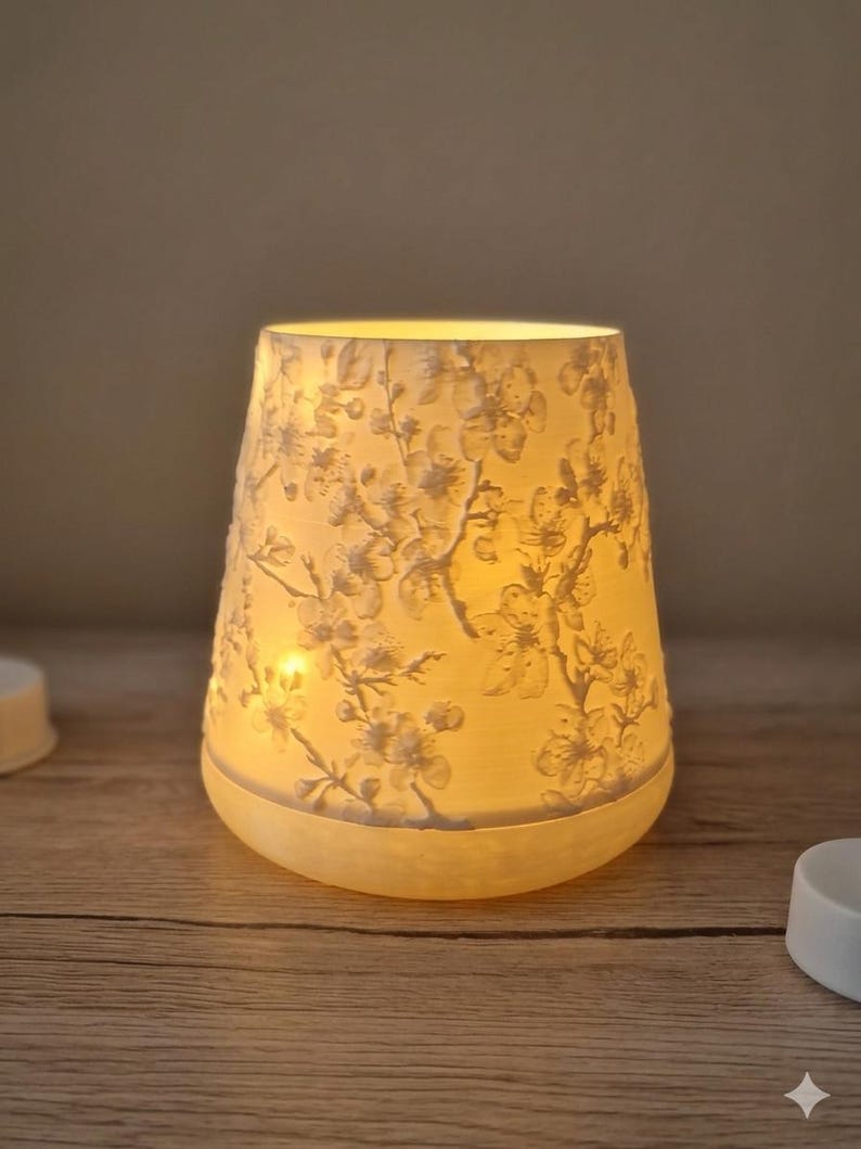 May include: A lit, cream-coloured candle holder with a floral pattern. The cylindrical holder has a flared top and a rounded base. The floral design is embossed on the surface, and the light from the candle inside illuminates the design. The candle holder sits on a wooden surface.