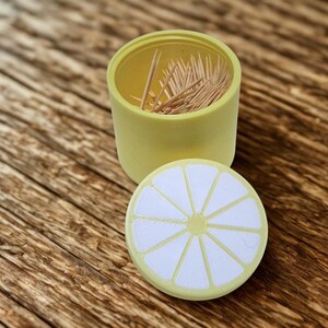 以下が含まれることがあります： 蓋にレモンスライスのデザインが施された黄色の楊枝入れ。容器は開いており、内部には木製の楊枝が見えます。蓋は丸く、白と黄色のレモンスライスのデザインです。