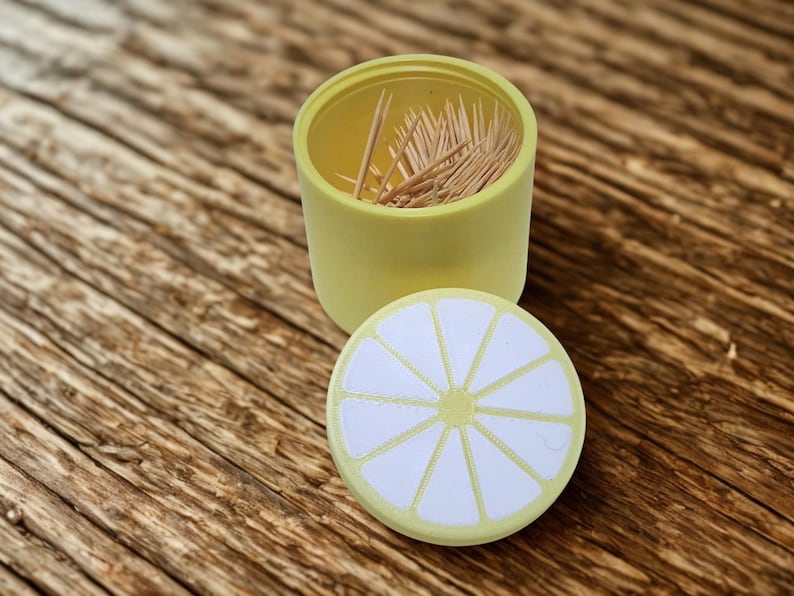 以下が含まれることがあります： 蓋にレモンスライスのデザインが施された黄色の楊枝入れ。容器は開いており、内部には木製の楊枝が見えます。蓋は丸く、白と黄色のレモンスライスのデザインです。
