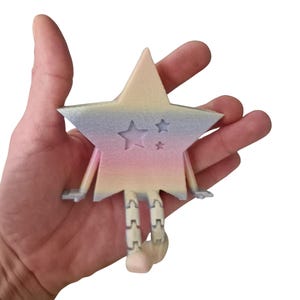 May include: A pastel-colored star-shaped figurine with a smiling face and star cutouts. The star has arms and segmented legs, and is held in a hand against a white background. The star's colors include pink, blue, and yellow.