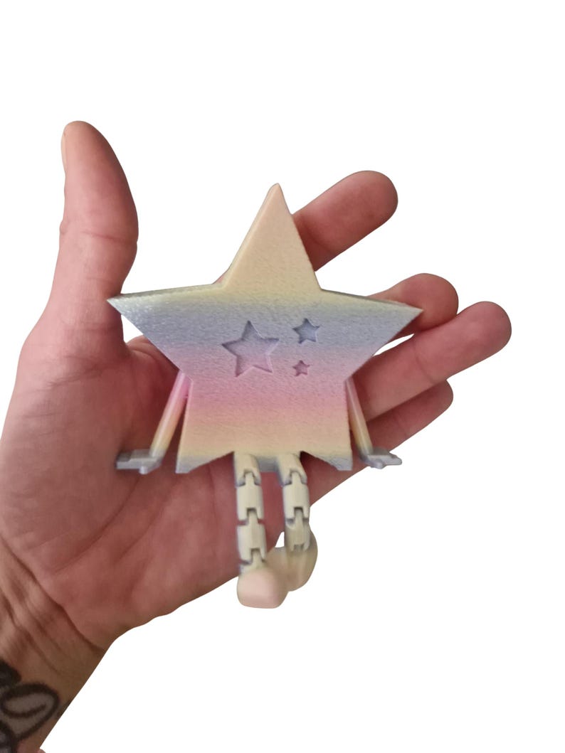 May include: A pastel-colored star-shaped figurine with a smiling face and star cutouts. The star has arms and segmented legs, and is held in a hand against a white background. The star's colors include pink, blue, and yellow.