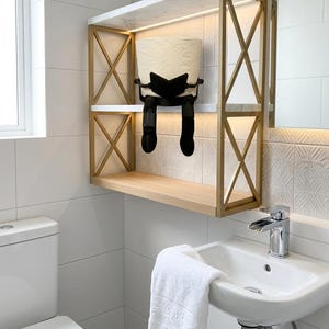 May include: A contemporary bathroom shelf with a gold metal frame and a white marble top. A black toilet paper holder shaped like a person is holding a roll of toilet paper. A white sink and towel are also visible.
