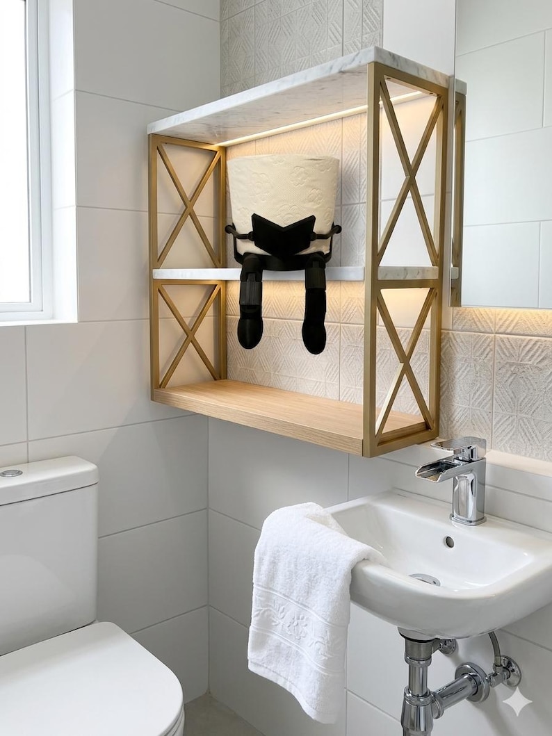 May include: A contemporary bathroom shelf with a gold metal frame and a white marble top. A black toilet paper holder shaped like a person is holding a roll of toilet paper. A white sink and towel are also visible.