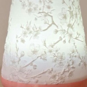 May include: A white, cylindrical lamp with a peach-coloured base. The lamp's surface features a delicate floral pattern of branches and blossoms. The design is raised, creating a textured effect. The lamp is lit from within, giving a soft glow.