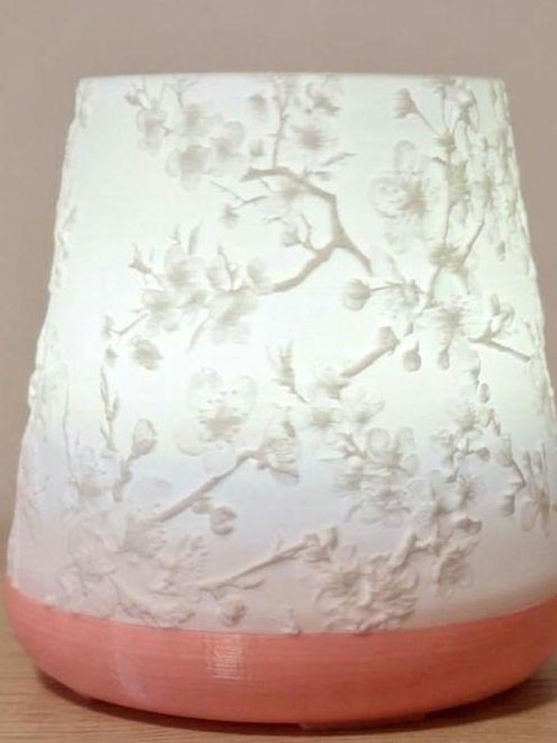 May include: A white, cylindrical lamp with a peach-coloured base. The lamp's surface features a delicate floral pattern of branches and blossoms. The design is raised, creating a textured effect. The lamp is lit from within, giving a soft glow.