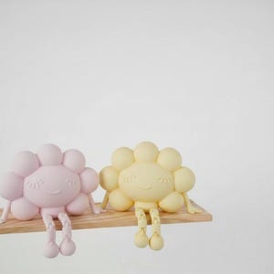 K&ouml;nnte beinhalten: Zwei fr&ouml;hliche, blumenf&ouml;rmige Figuren, eine rosa und eine gelbe, sitzen auf einem kleinen Holzregal. Jede Figur hat einen runden Kopf mit bl&uuml;tenblattartigen Formen und h&auml;ngenden Beinen. Der Hintergrund ist wei&szlig;.
