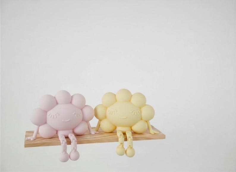 K&ouml;nnte beinhalten: Zwei fr&ouml;hliche, blumenf&ouml;rmige Figuren, eine rosa und eine gelbe, sitzen auf einem kleinen Holzregal. Jede Figur hat einen runden Kopf mit bl&uuml;tenblattartigen Formen und h&auml;ngenden Beinen. Der Hintergrund ist wei&szlig;.