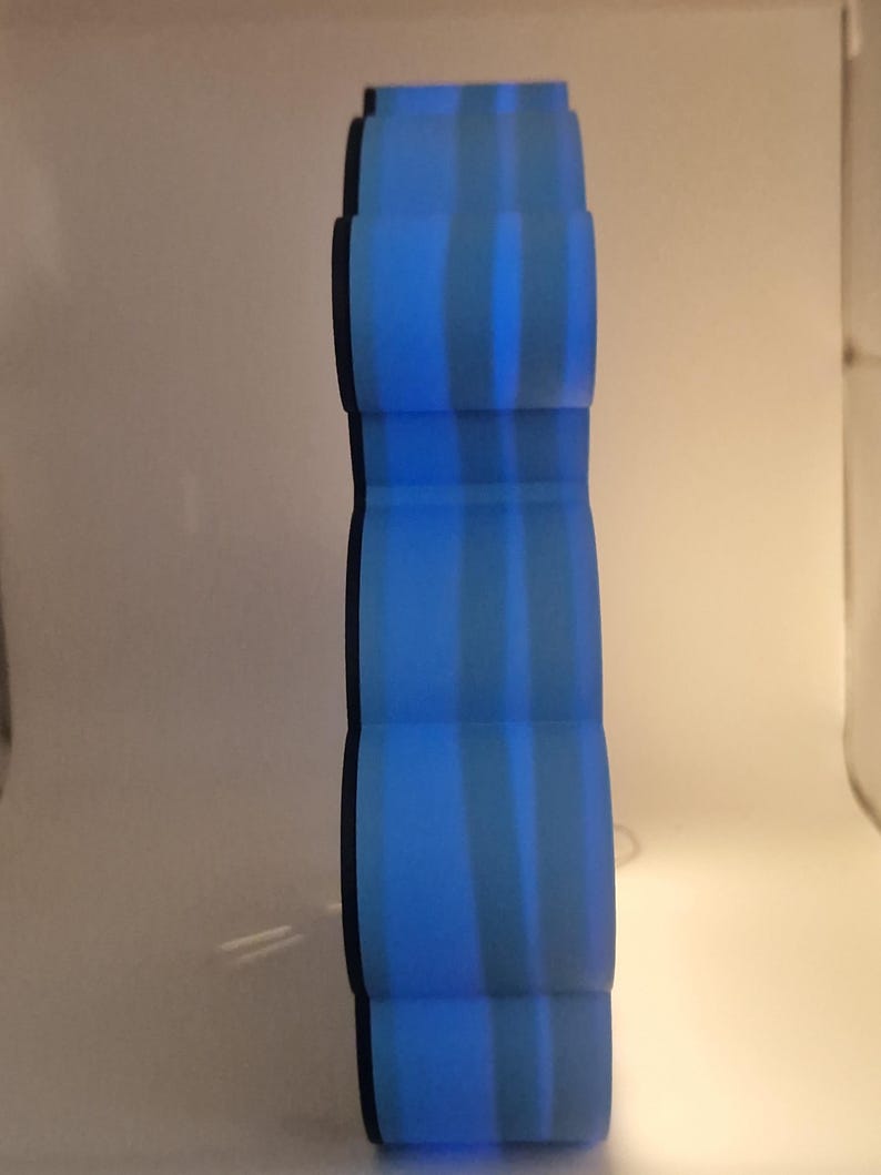 Puede incluir: Un jarr&oacute;n decorativo azul, alto y con un dise&ntilde;o &uacute;nico en capas. El jarr&oacute;n tiene forma cil&iacute;ndrica con secciones redondeadas apiladas. Rayas verticales de color azul claro crean un patr&oacute;n a rayas. El jarr&oacute;n mide unos 30 cm de altura.