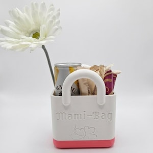 以下が含まれることがあります： 白いハンドルが付いた白とピンクの「Mami-Bag」トートバッグ。缶や包装されたアイテムが入っています。左側に白い花があります。バッグには「Mami-Bag」の文字とハートのデザインがあります。