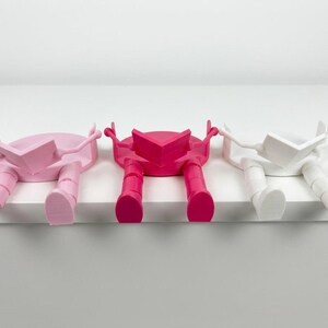 May include: Three colourful bookends, each in a different shade: pale pink, bright pink, and white. The bookends are designed as figures sitting and reading, with legs hanging over a shelf edge. Each figure is holding an open book.