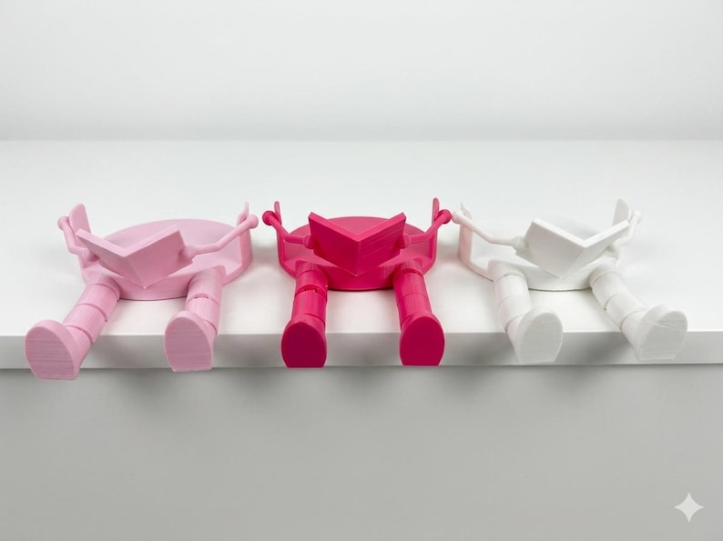 May include: Three colourful bookends, each in a different shade: pale pink, bright pink, and white. The bookends are designed as figures sitting and reading, with legs hanging over a shelf edge. Each figure is holding an open book.