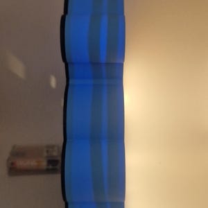 Puede incluir: Un objeto cil&iacute;ndrico azul con rayas verticales. El objeto tiene una serie de secciones redondeadas apiladas. El fondo es de color neutro. Un peque&ntilde;o objeto rectangular es visible en el fondo.