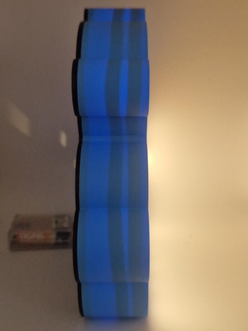 Puede incluir: Un objeto cil&iacute;ndrico azul con rayas verticales. El objeto tiene una serie de secciones redondeadas apiladas. El fondo es de color neutro. Un peque&ntilde;o objeto rectangular es visible en el fondo.