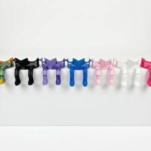 May include: A collection of colourful bookends, each designed as a person reading. The bookends are in a range of colours: rainbow, black, purple, blue, pink, white, and red. They are arranged on a white surface.