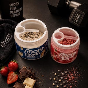 Könnte beinhalten: Zwei offene Behälter mit More Chunky Flavour Proteinpulver, einer mit blauem und einer mit rosa Etikett. Ein schwarzes Hantel, Erdbeeren und Schokolade sind ebenfalls zu sehen.