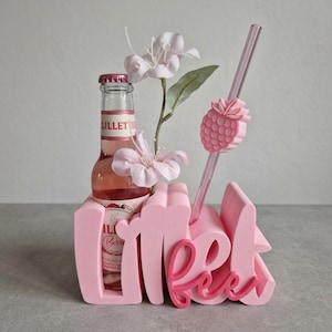 Pode incluir: Um suporte rosa decorativo para bebidas com a palavra "Meet" em uma fonte l&uacute;dica. Uma garrafa de Lillet Berry est&aacute; colocada dentro, com um canudo rosa e um detalhe em forma de abacaxi. Flores artificiais adicionam um toque de eleg&acirc;ncia.