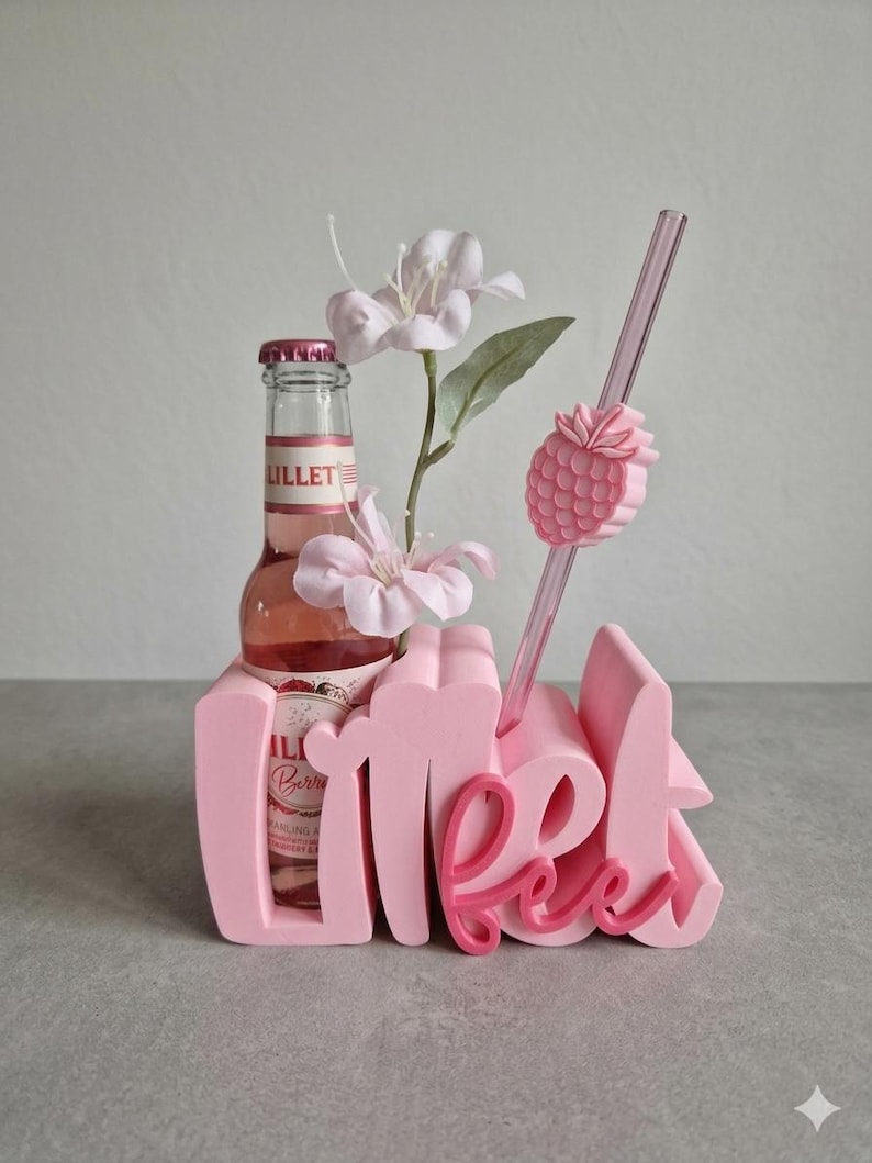 Pode incluir: Um suporte rosa decorativo para bebidas com a palavra "Meet" em uma fonte l&uacute;dica. Uma garrafa de Lillet Berry est&aacute; colocada dentro, com um canudo rosa e um detalhe em forma de abacaxi. Flores artificiais adicionam um toque de eleg&acirc;ncia.