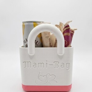 以下が含まれることがあります： ピンクのベースと白いハンドルが付いた白いトートバッグで、「Mami-Bag」と表示されています。バッグには缶といくつかの包装されたアイテムが入っています。テキストの下にはハート型のデザインがあります。バッグは小さなアイテムを収納するように設計されています。
