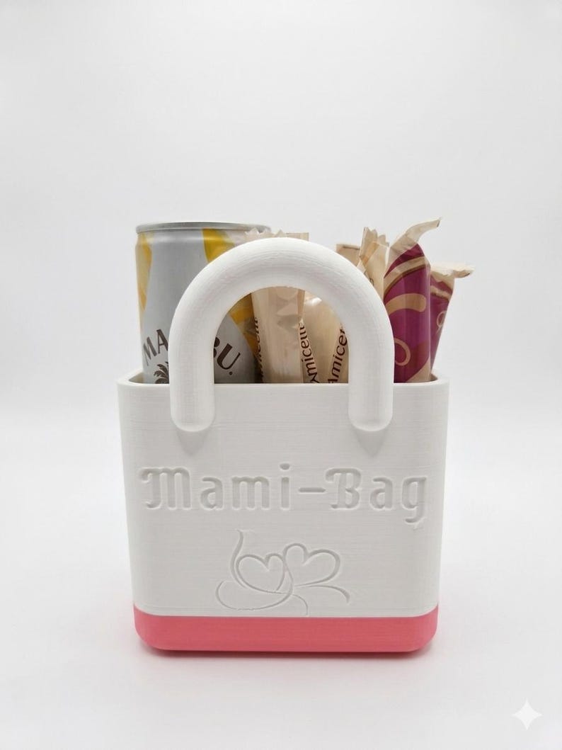 以下が含まれることがあります： ピンクのベースと白いハンドルが付いた白いトートバッグで、「Mami-Bag」と表示されています。バッグには缶といくつかの包装されたアイテムが入っています。テキストの下にはハート型のデザインがあります。バッグは小さなアイテムを収納するように設計されています。